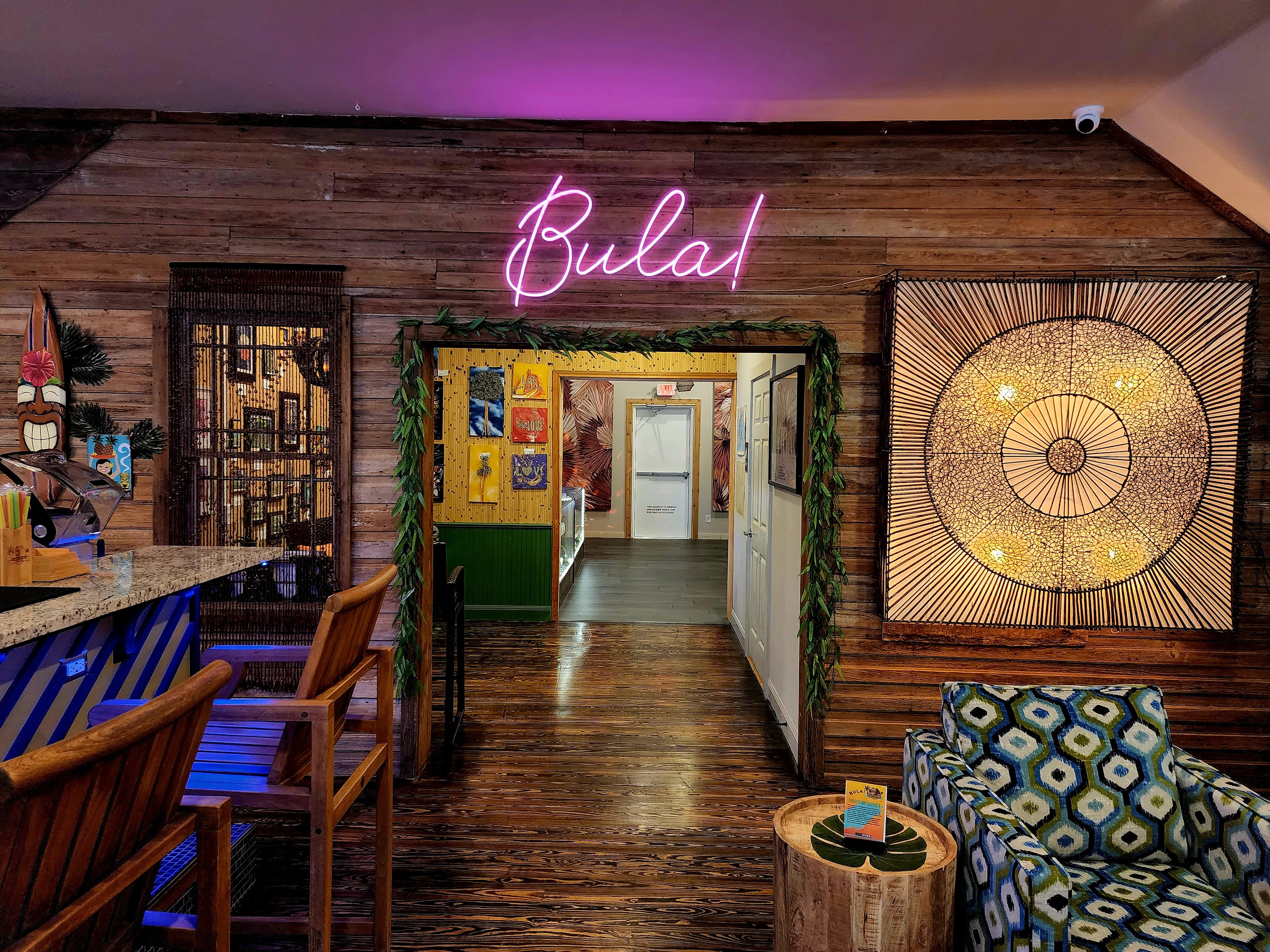 Bula neon glowing over the entryway with tiki mask and woven sunburst art inside Elixir Lounge.