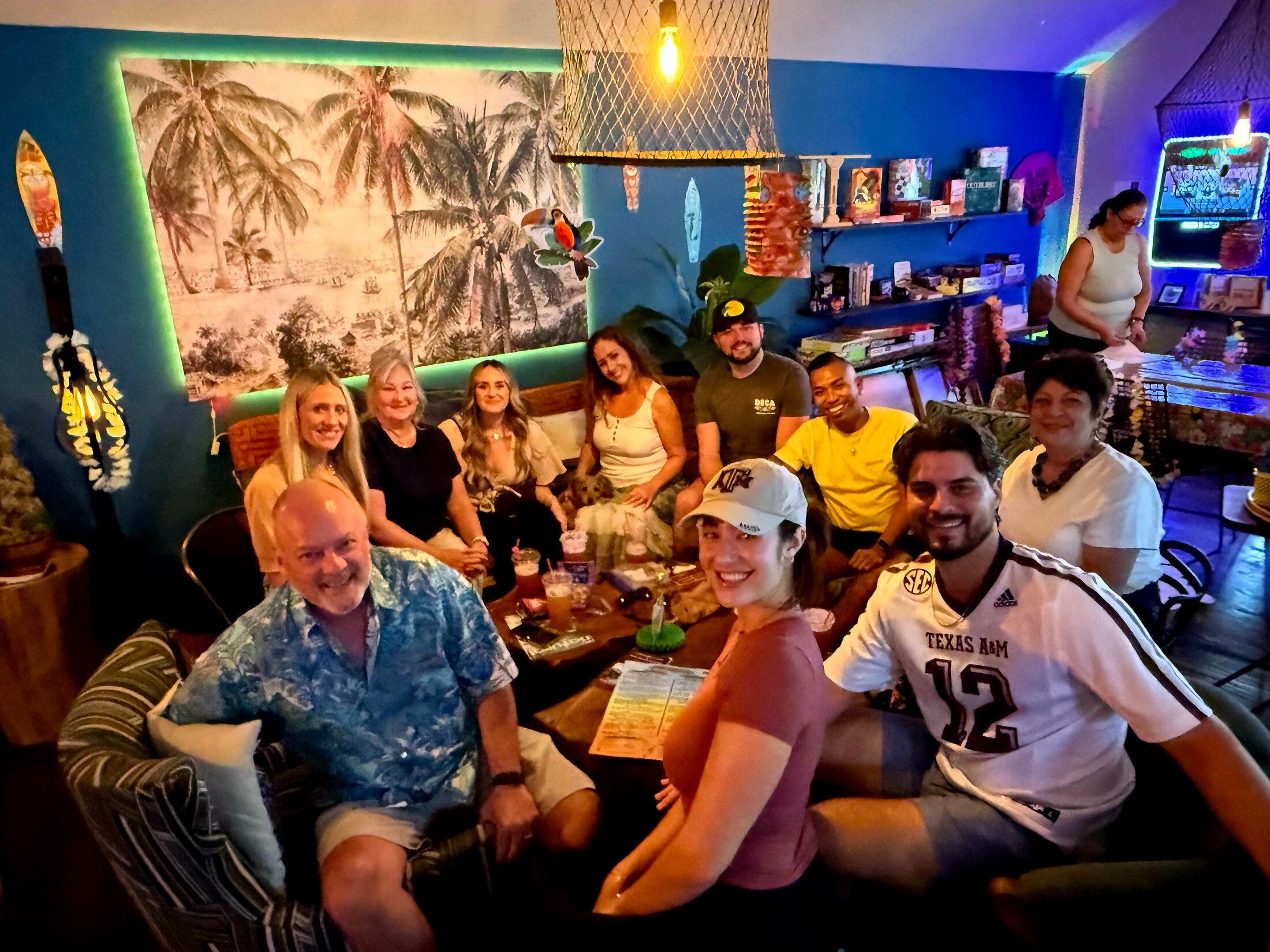Smiling group of guests gathered on couches under a tropical palm mural at Elixir Lounge.