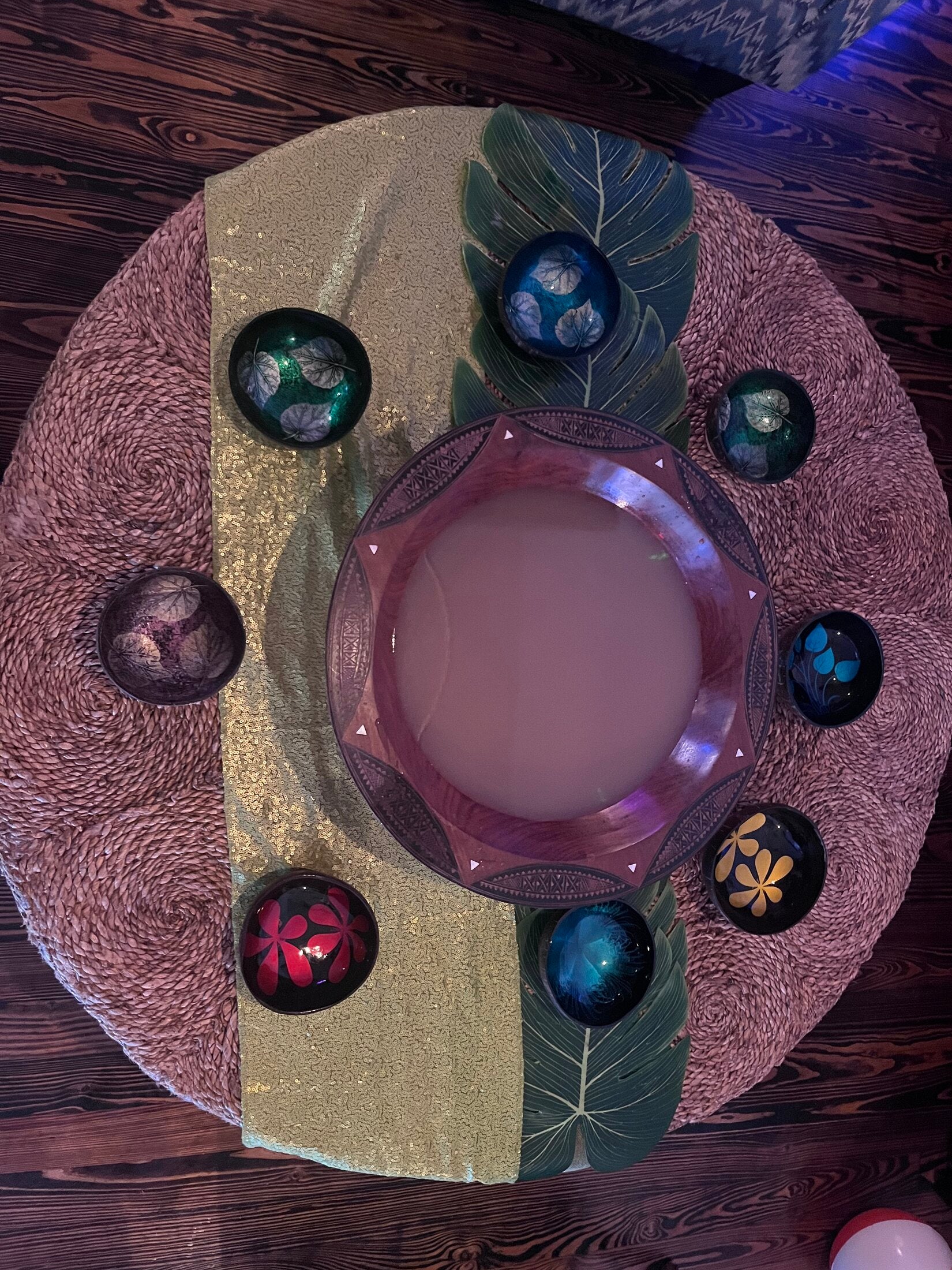 Overhead view of a carved kava tanoa bowl ringed by decorated coconut shell cups on a woven mat at Elixir Lounge.