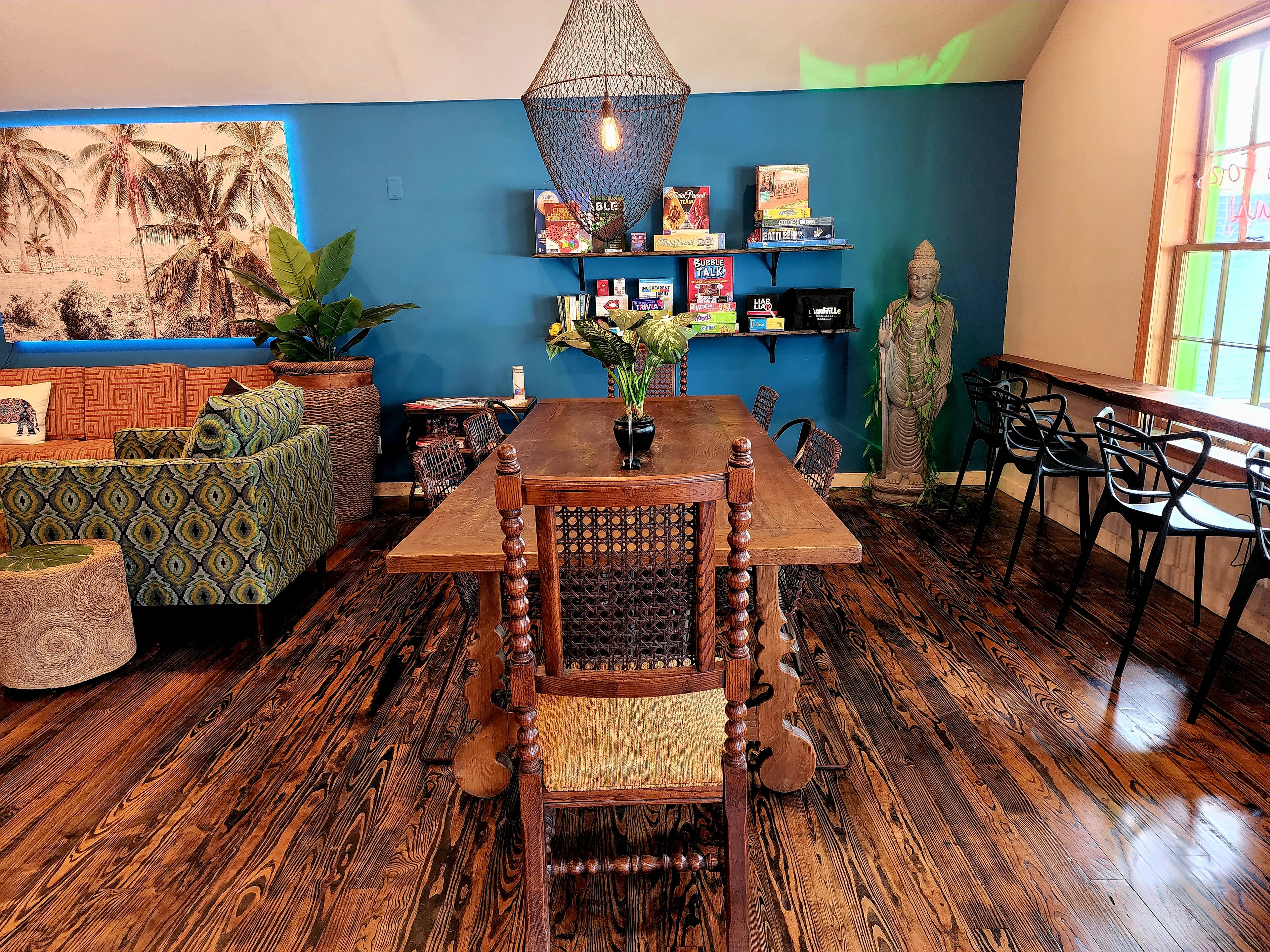 Blue-wall lounge area with a communal table, board-game shelf and standing Buddha statue at Elixir Lounge.