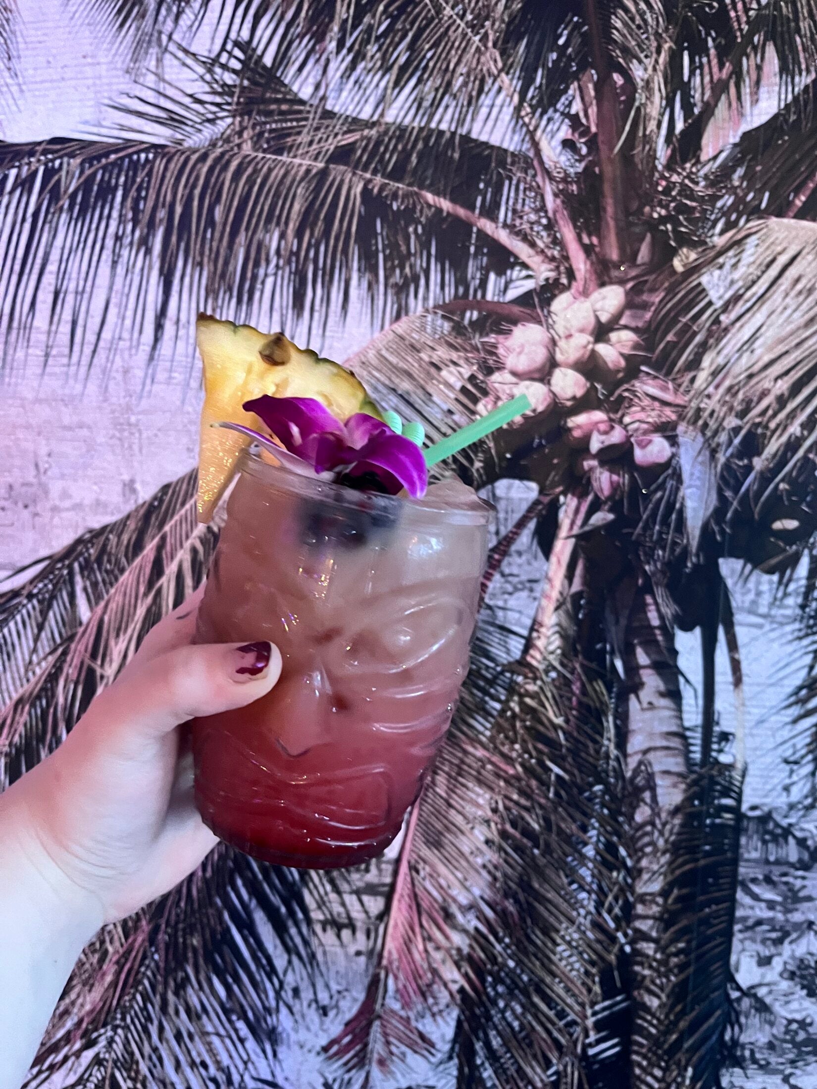 Tropical drink garnished with pineapple and a purple orchid against a palm-tree mural at Elixir Lounge.