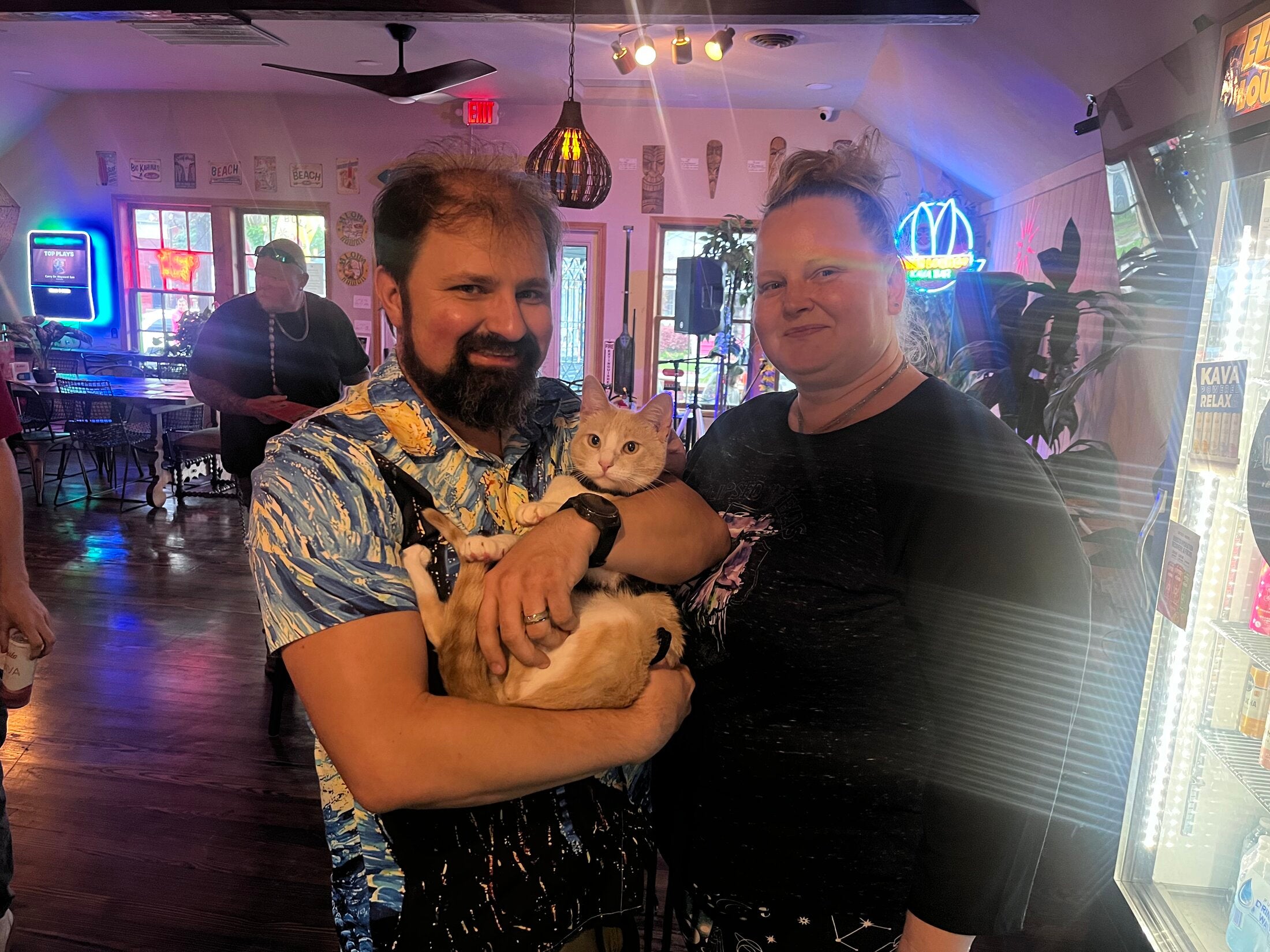 Smiling couple holding their cat inside the pet-friendly Elixir Lounge with tiki wall decor.
