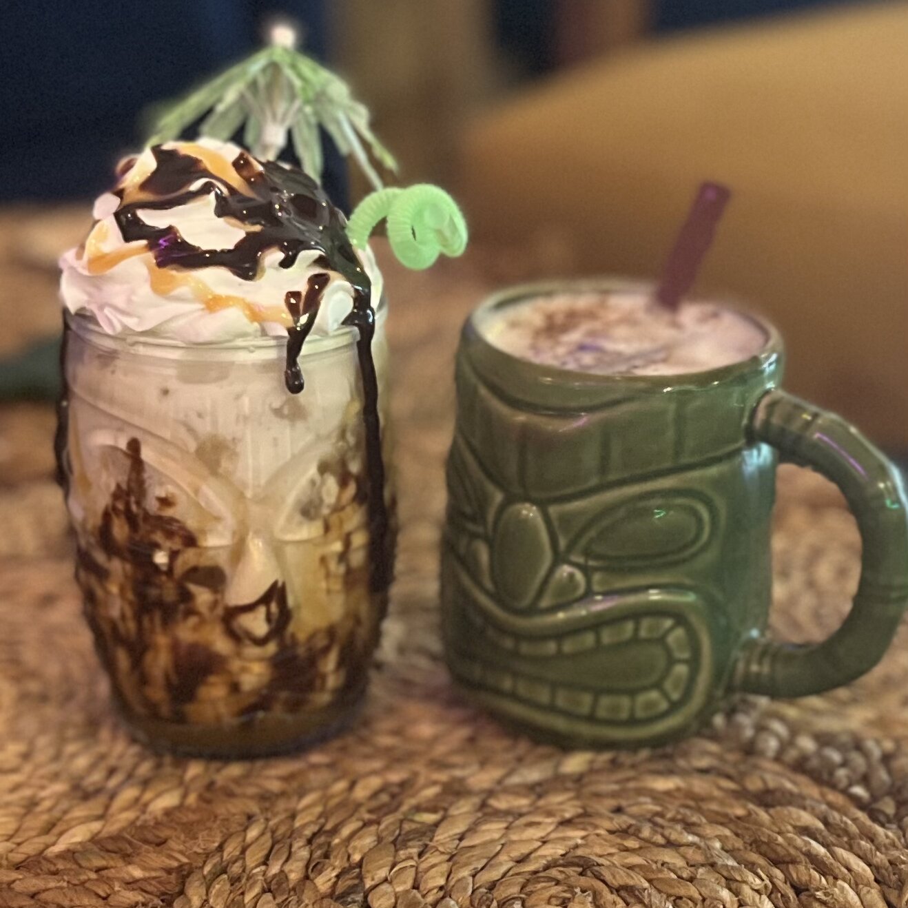 Whipped-cream mason-jar drink and a green tiki mug drink side by side on a woven mat at Elixir Lounge.