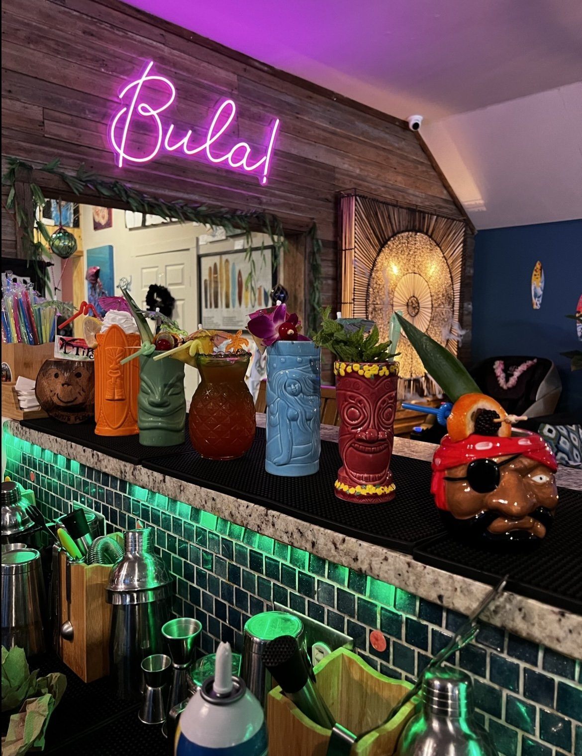 Lineup of colorful tiki mugs with garnished drinks on the bar under the Bula! neon