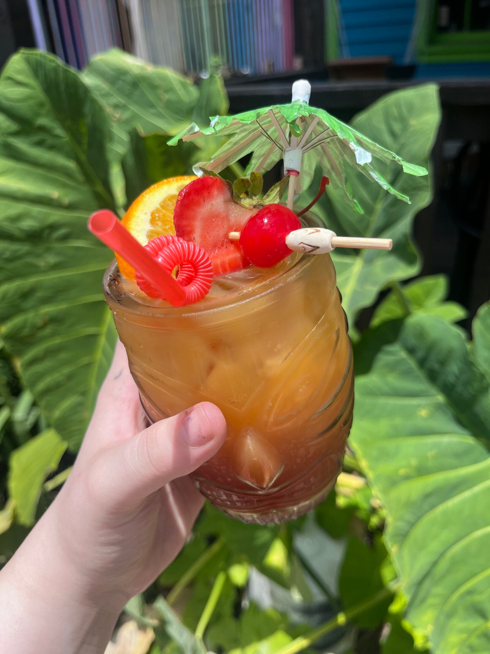 Amber tiki-mug drink with orange, strawberry, cherry and umbrella garnish against tropical leaves.